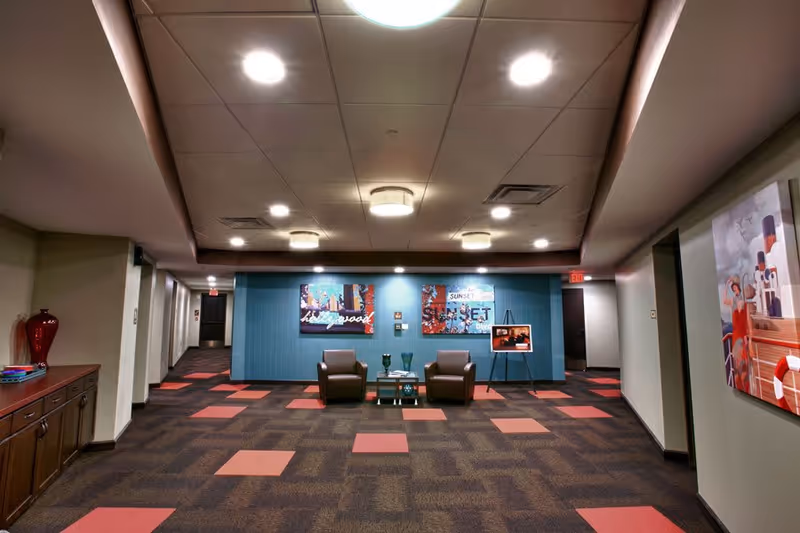 A spacious interior hallway area with patterned carpet featuring red and brown squares. Two brown armchairs and a small table with decorative items are positioned against a blue accent wall with colorful artwork. The ceiling has multiple recessed lights and fixtures. There is a wooden cabinet with a red vase on the left side and a painting on the right wall.