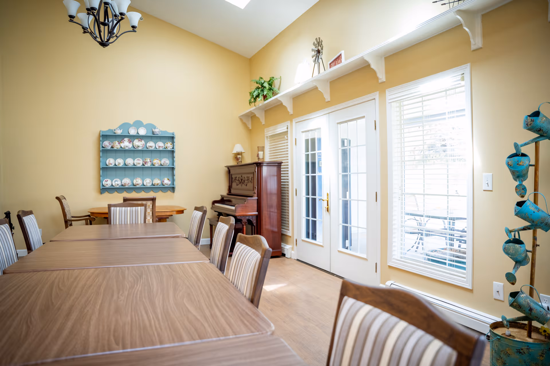 Bright communal dining room with long tables and chairs, a piano, decorative plate rack, and French doors leading outside.