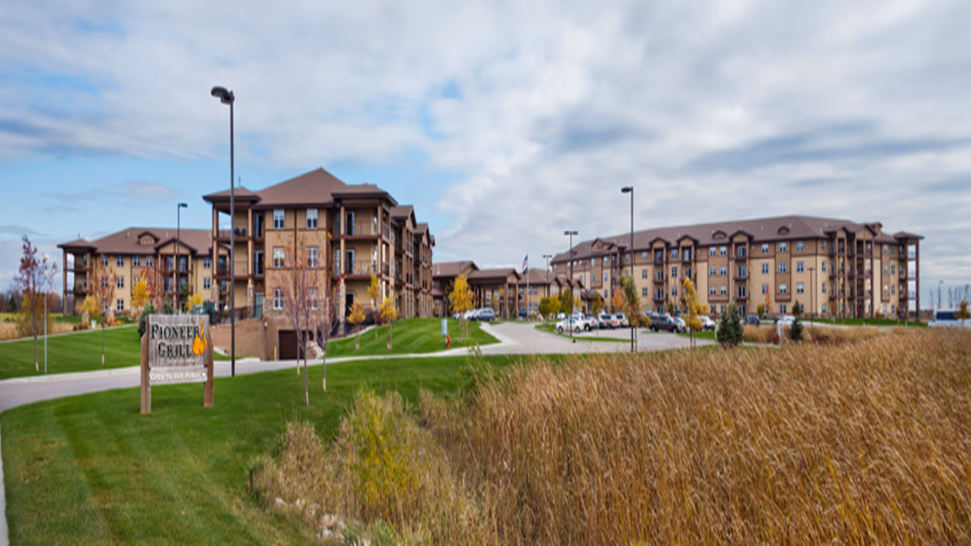 Exterior view of a large senior living facility with multiple connected buildings, surrounded by green lawns and tall grass. There is a sign in the foreground that reads 'Pioneer Grill Open to the Public'. Several cars are parked near the buildings under a partly cloudy sky.