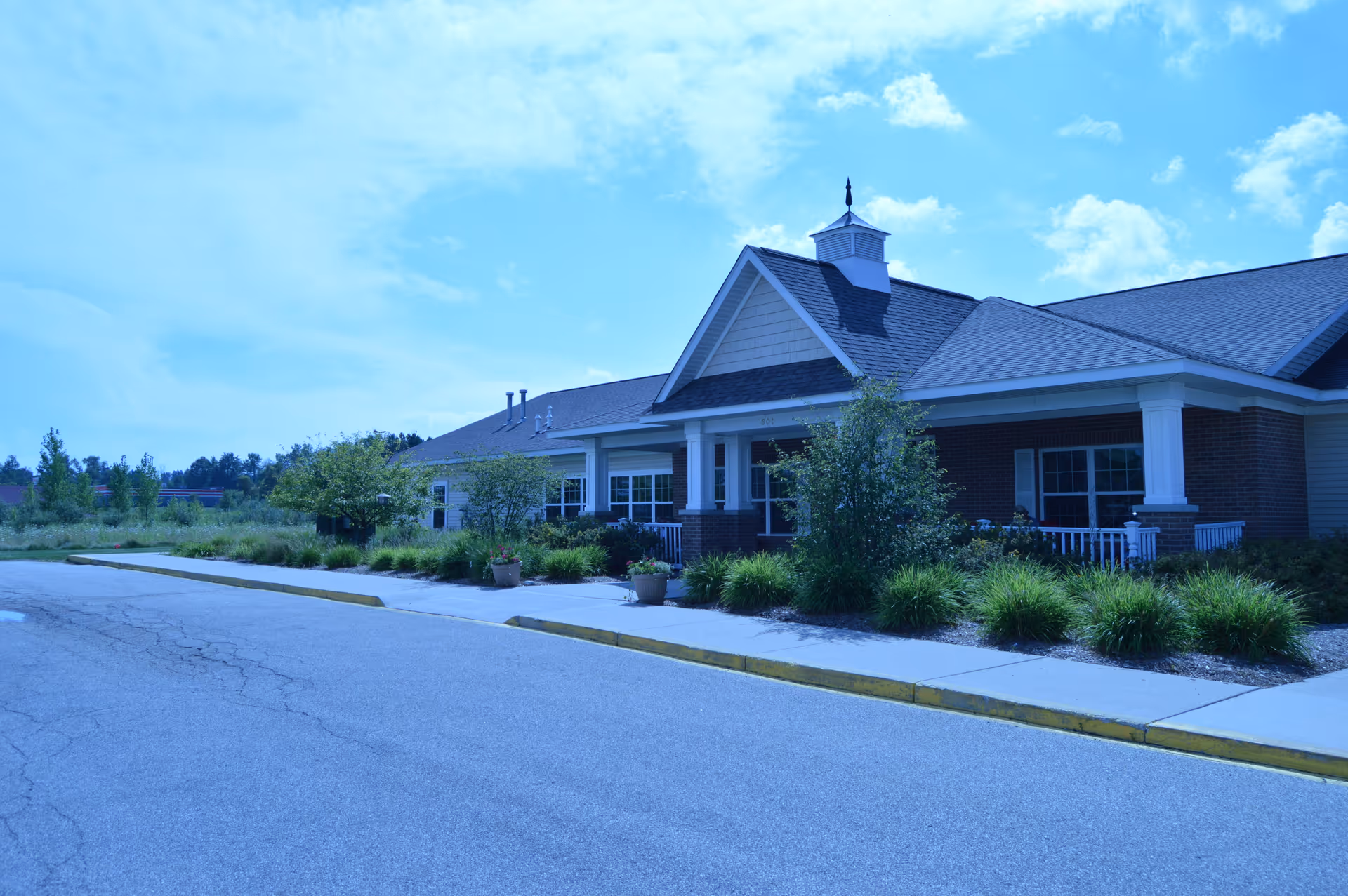 Single-story brick assisted living building with a covered porch entrance, surrounding shrubs, and an empty driveway.