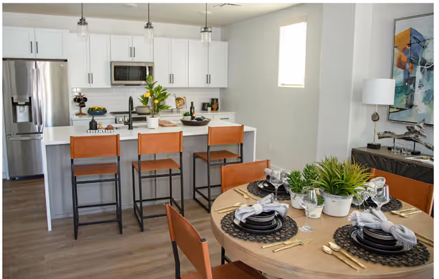 Modern kitchen and dining area with white cabinets, stainless steel refrigerator, and a kitchen island with three brown leather bar stools. A round dining table is set with black plates, gold cutlery, gray napkins, and decorative green plants. The room has light wood flooring and a window with a white blind.