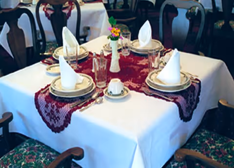 A dining table set for four with white tablecloth and a burgundy lace table runner. Each place setting includes a plate, a folded white napkin, a glass, and silverware. A small vase with flowers is in the center of the table. The chairs around the table have floral patterned cushions.