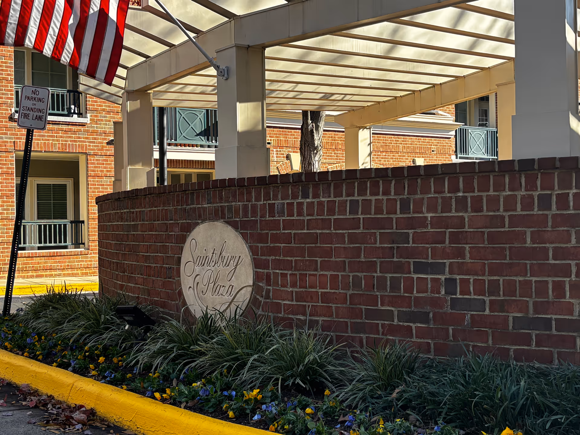 Brick entrance wall with a round "Saintsbury Plaza" plaque, landscaping, an American flag, and a covered pergola at the front of the building.