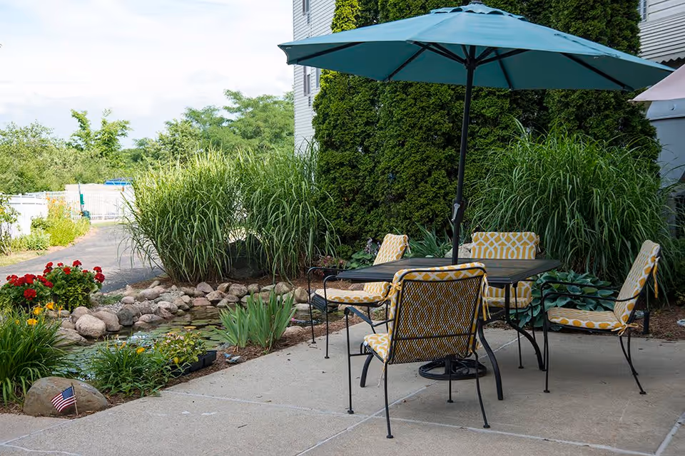Outdoor patio area with a table and four yellow patterned cushioned chairs under a large green umbrella, surrounded by lush greenery, plants, and a small pond with rocks and flowers.