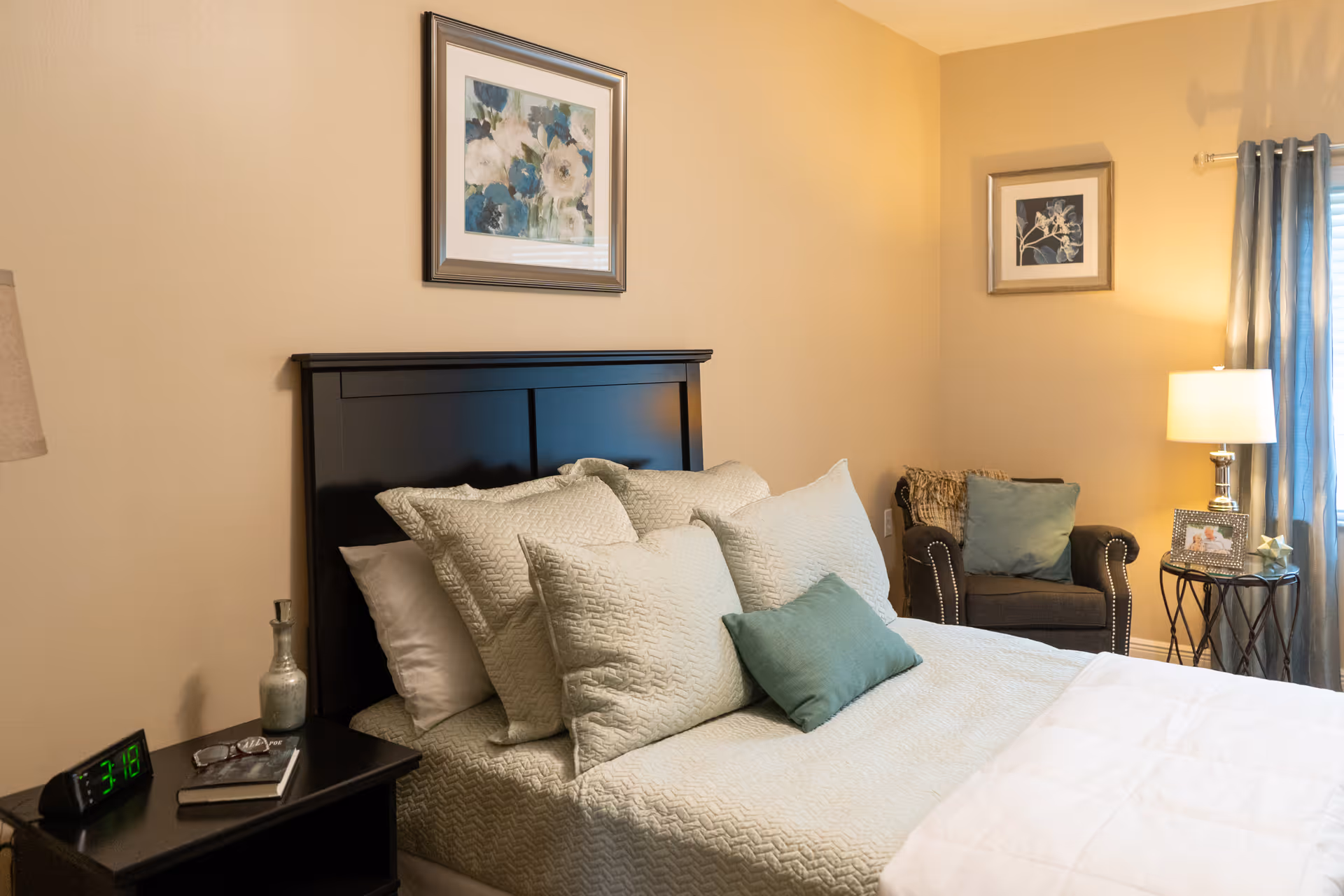 A cozy bedroom featuring a neatly made bed with multiple pillows in light green and white shades. The bed has a dark wooden headboard. Next to the bed is a black nightstand with a digital clock showing 3:18, a pair of glasses, a book, and a decorative bottle. In the corner, there is a comfortable armchair with a green cushion and a throw blanket. A small round side table beside the chair holds a lamp and a framed photo. The walls are painted beige and decorated with framed floral artwork. A window with blue curtains allows natural light into the room.