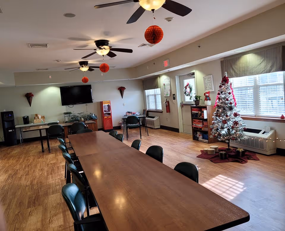 A spacious common room with a long wooden table surrounded by chairs. The room has three ceiling fans with lights, red hanging decorations, a white Christmas tree with red ornaments and presents underneath, a wall-mounted TV, a water cooler, and a small bookshelf with board games. Large windows let in natural light, and there is a door with a wreath on it.