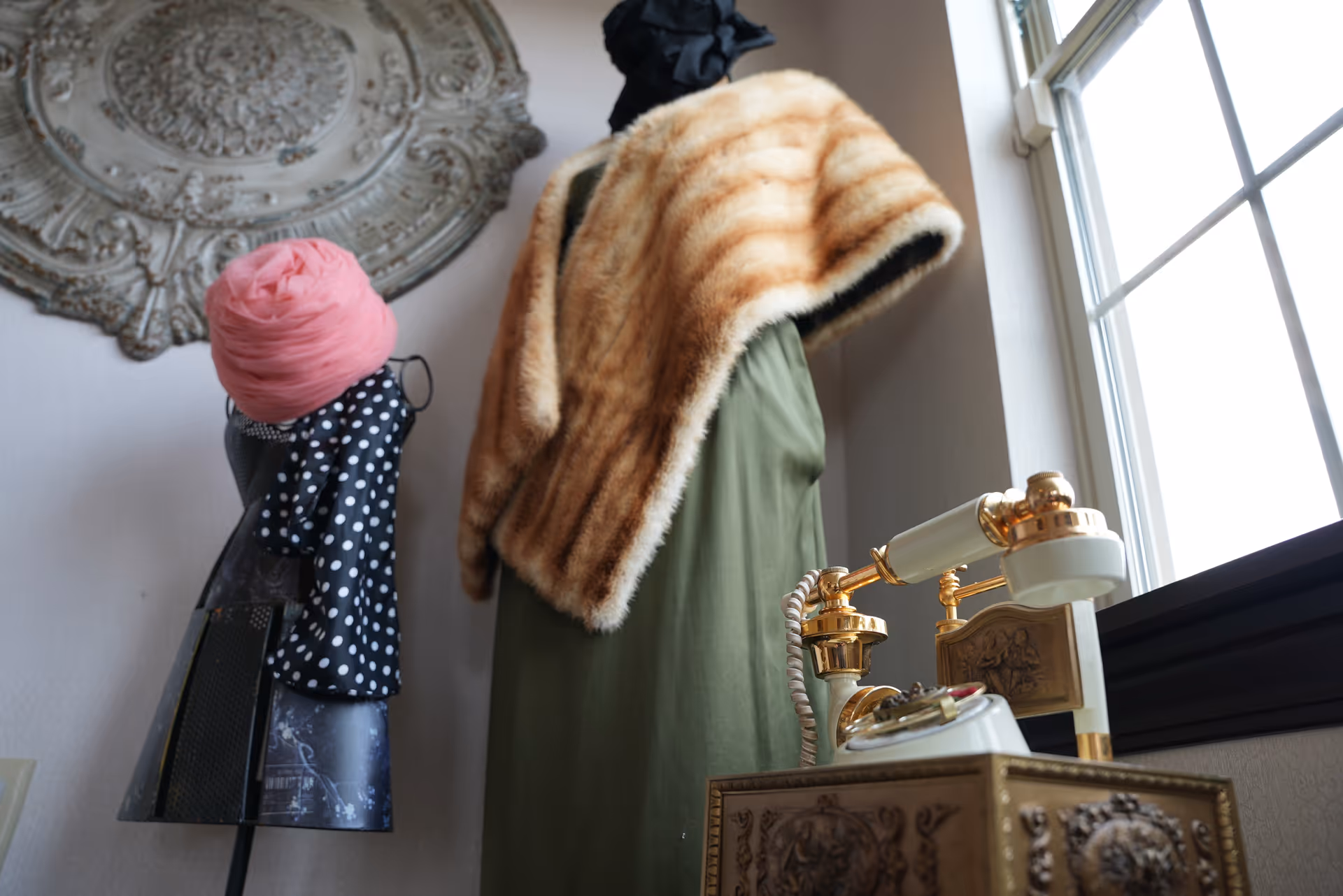 A decorative corner of a room showing a dress form draped in a fur stole, a vintage telephone on an ornate box, and a window.