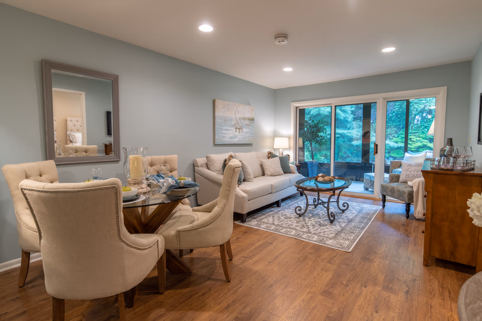 A cozy living room and dining area with light beige upholstered chairs around a glass dining table, a beige sofa with decorative pillows, a round glass coffee table on a patterned rug, and a sliding glass door leading to a green outdoor space. The walls are painted light blue, and there is a large mirror and a painting on the walls.