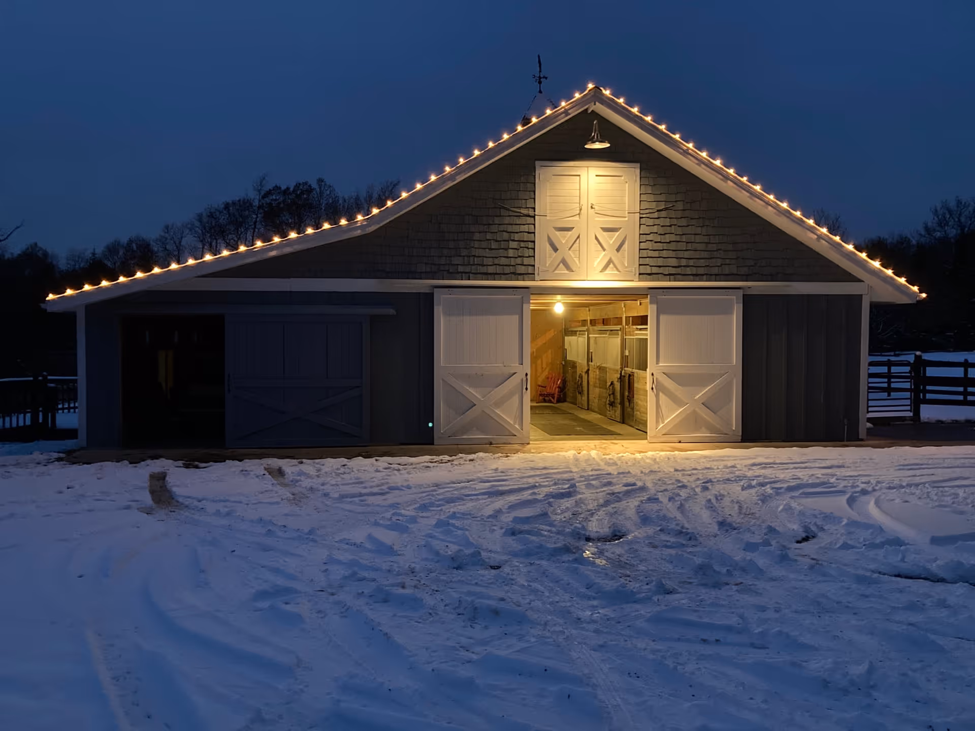A barn-style building lit by string lights with open central doors and snow-covered ground in front at dusk.