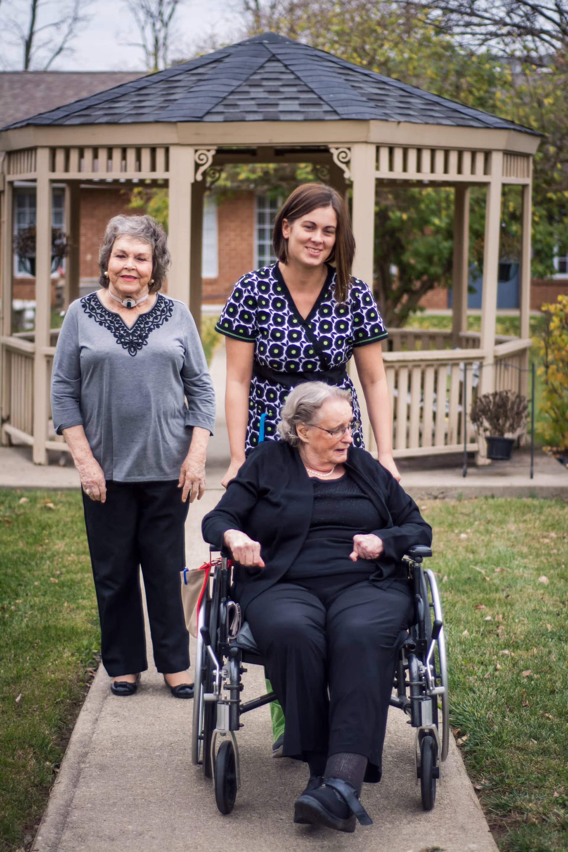 An elderly woman in a wheelchair is being pushed by a younger woman along a paved path in a garden area with a gazebo in the background. Another elderly woman is walking beside them, smiling.
