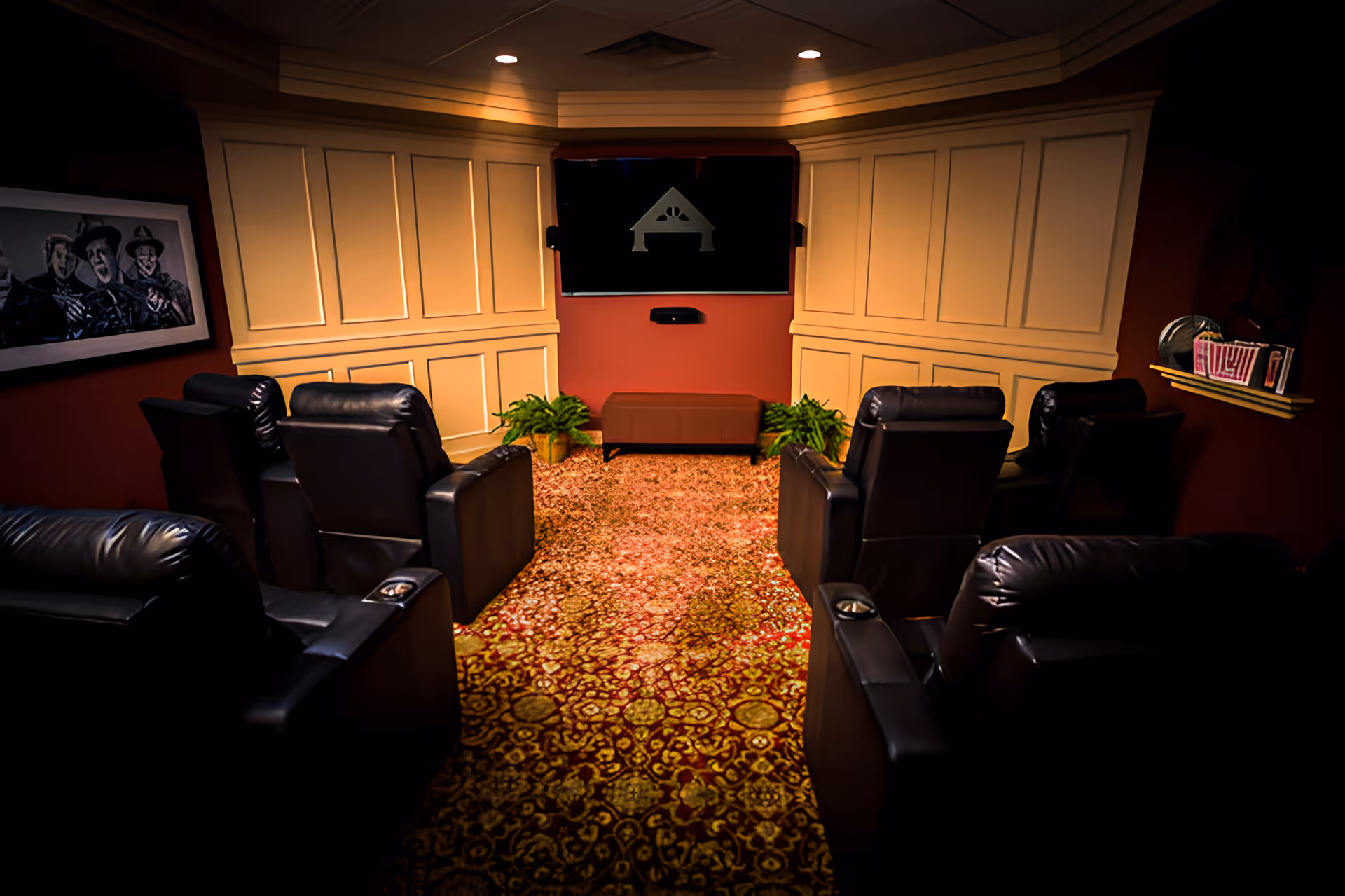 A small home theater room with six black leather recliner chairs arranged in two rows facing a wall-mounted flat screen TV. The room has beige paneled walls, a patterned carpet with red and gold tones, two green potted plants on either side of a brown ottoman below the TV, and a framed black and white photo on the left wall.