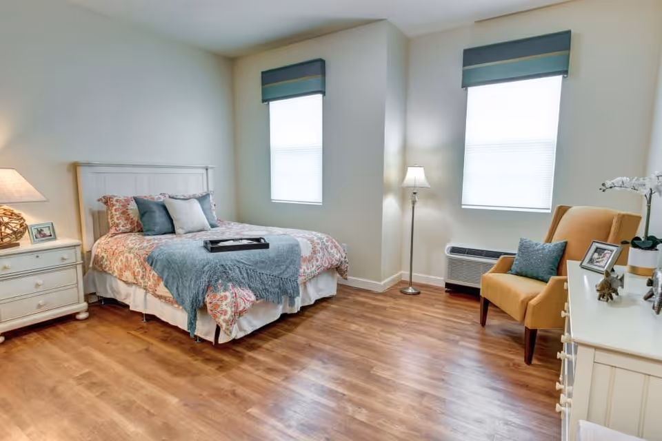A bright and cozy bedroom with a bed featuring floral bedding and multiple pillows, a nightstand with a lamp and framed photo, two windows with blue valances, a floor lamp, a mustard yellow armchair with a blue cushion, and a white dresser with decorative items including a small plant and framed photo. The room has wooden flooring and light-colored walls.