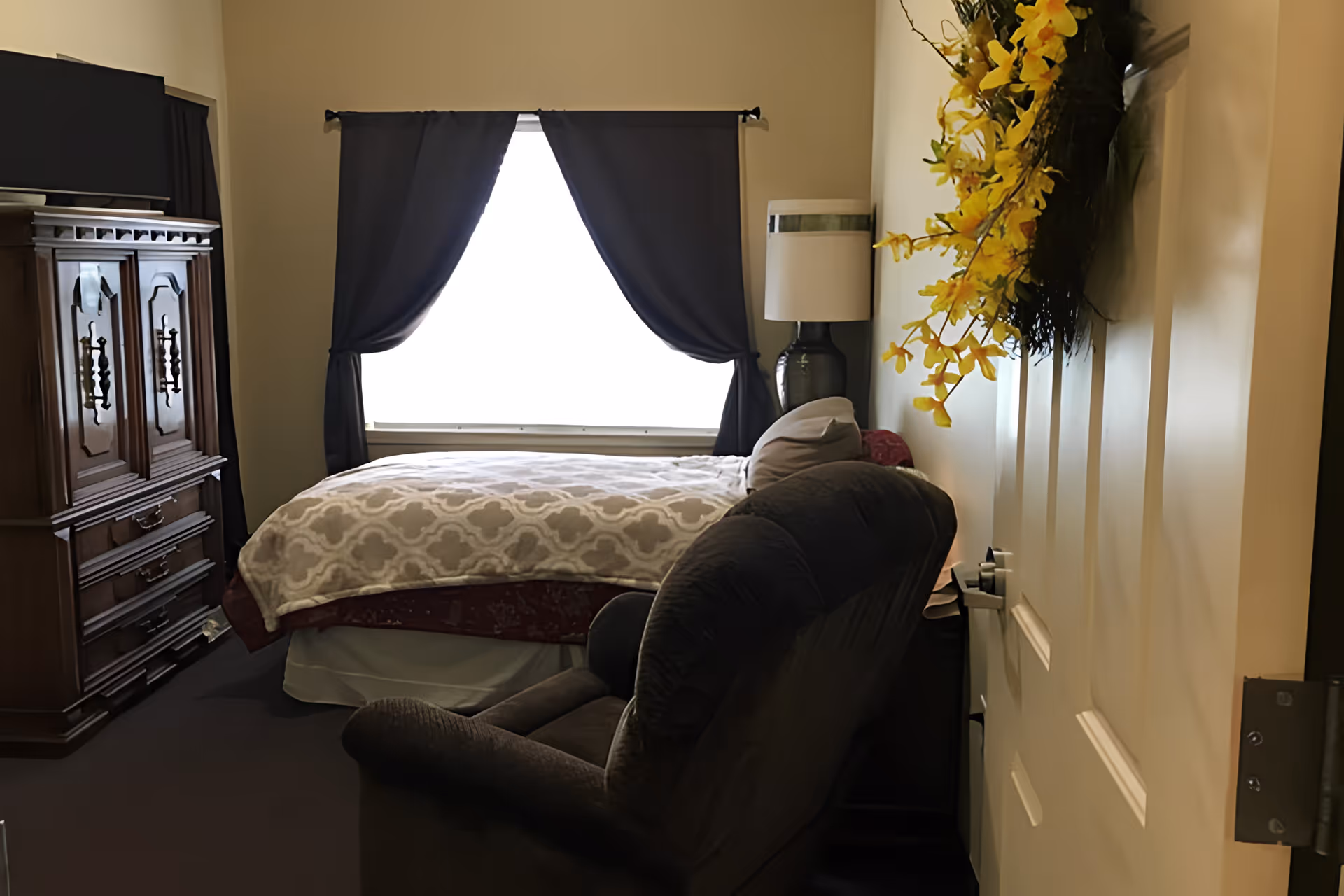 A cozy bedroom with a bed covered in a patterned blanket positioned against a window with dark curtains. To the left, there is a wooden armoire with a television on top. A cushioned armchair is placed near the foot of the bed, and a lamp with a white shade sits on a nightstand beside the bed. A decorative wreath with yellow flowers hangs on the door.