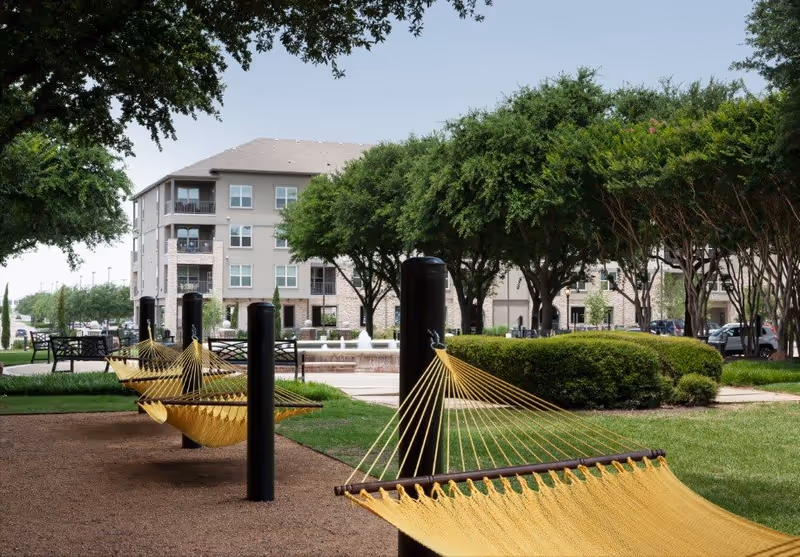Outdoor area with yellow hammocks suspended between black posts, surrounded by green grass, trees, and bushes. In the background, there is a multi-story residential building and a water fountain.