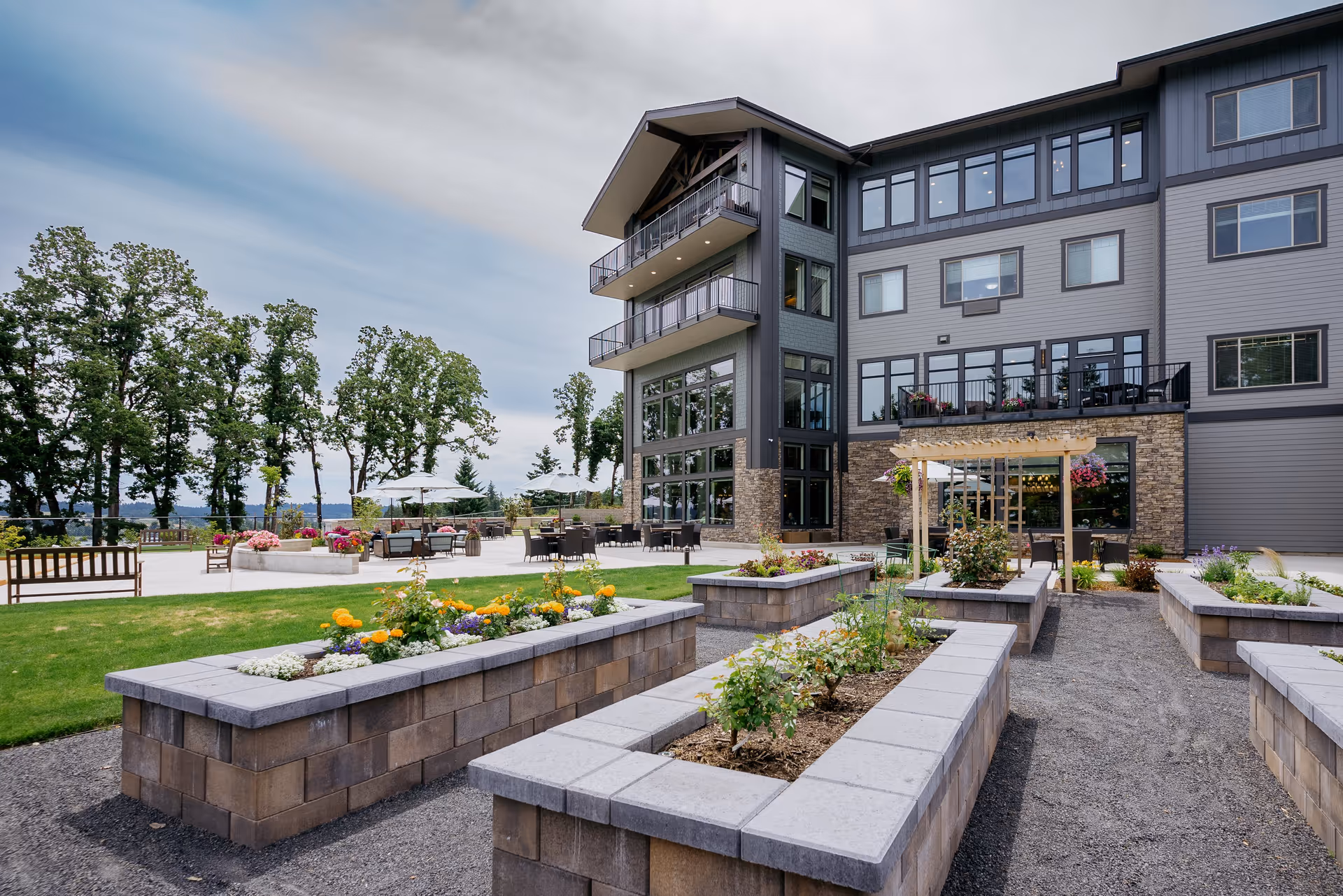 Outdoor garden area with raised flower beds containing various plants and flowers in front of a multi-story retirement community building. The building has large windows and balconies. There are patio tables with umbrellas and chairs on a paved area, surrounded by green grass and trees under a cloudy sky.