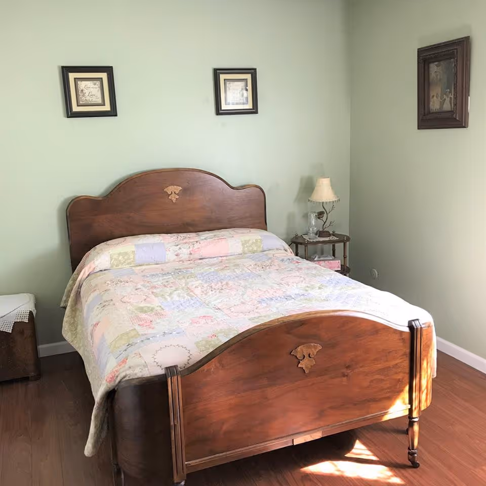 A bedroom with a wooden bed frame and a patchwork quilt on the bed. The walls are painted light green and decorated with three framed pictures. There is a small wooden side table with a lamp and a glass base next to the bed, and a wooden chest with a white lace cover in the corner.