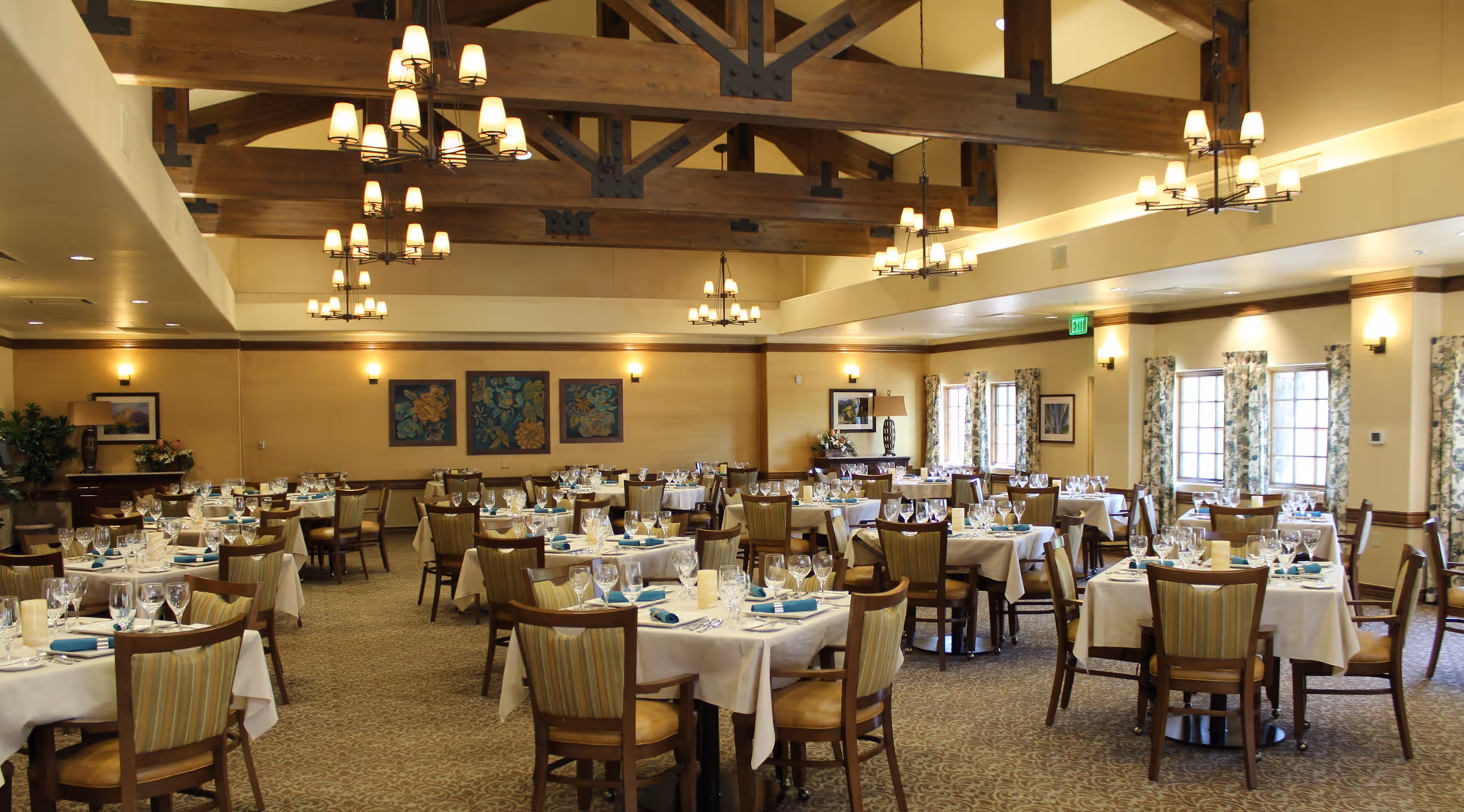 A spacious dining room with multiple round tables covered with white tablecloths, each set with glassware, plates, and blue napkins. The room features wooden beams on the ceiling, several chandeliers, framed floral artwork on the walls, and windows with floral curtains allowing natural light to enter.
