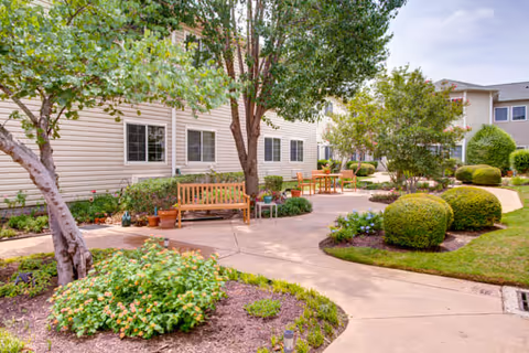 Outdoor courtyard area at The Court at Round Rock featuring a paved walkway, wooden benches, green shrubs, trees, and a well-maintained garden with flowering plants, adjacent to a beige residential building.