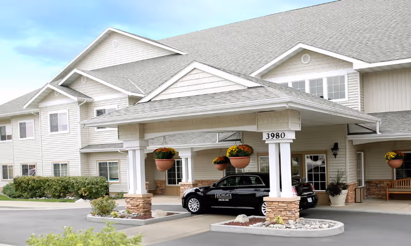 Exterior view of Highgate Senior Living facility showing the main entrance with a covered drop-off area supported by white columns with stone bases. A black car with Highgate Senior Living branding is parked under the entrance canopy. The building is two stories with beige siding, multiple windows, and a gray shingled roof. Hanging flower pots and landscaping with bushes and rocks are visible around the entrance.