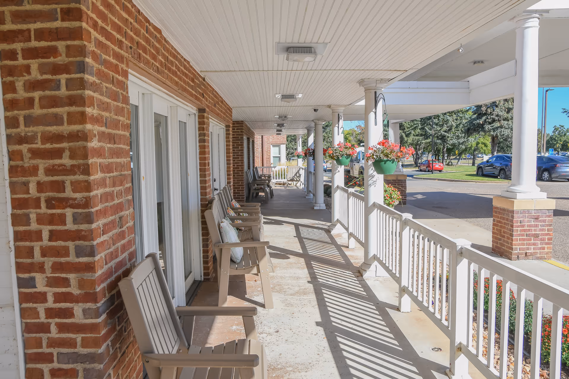 A covered outdoor porch area with several beige chairs lined up against a brick wall. White columns support the roof, and hanging flower pots with red and pink flowers are attached to the columns. A white railing runs along the edge of the porch, and a parking lot with cars and trees is visible in the background under a clear blue sky.