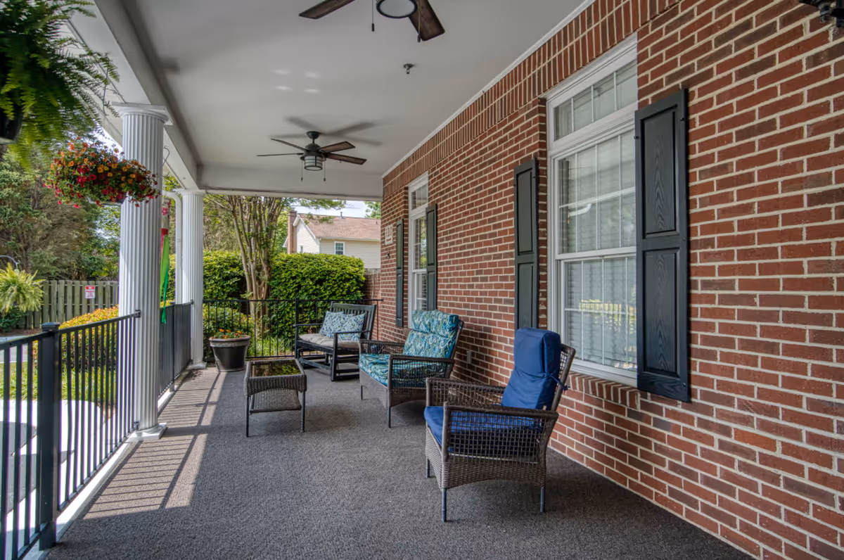 Covered front porch with wicker chairs, potted plants, and ceiling fans along a brick building.