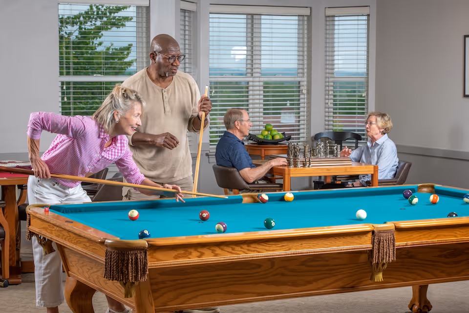 Four people socialize in a bright game room with a pool table in the foreground and a chess game at a table in the background.