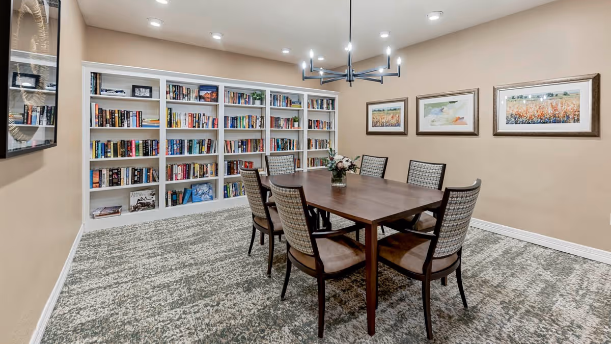 Community reading room with a wooden table surrounded by six chairs and a wall of bookshelves.