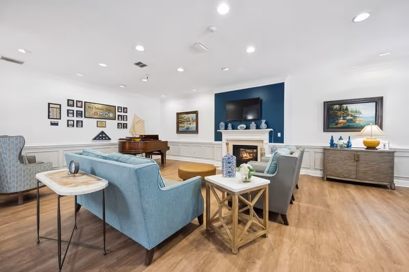 A bright and spacious living room with light blue and gray armchairs and sofas arranged around a white fireplace with a TV mounted above it. The room features wooden flooring, a grand piano, framed artwork on the walls, and decorative items on side tables and a console. The walls are white with wainscoting, and the ceiling has recessed lighting.