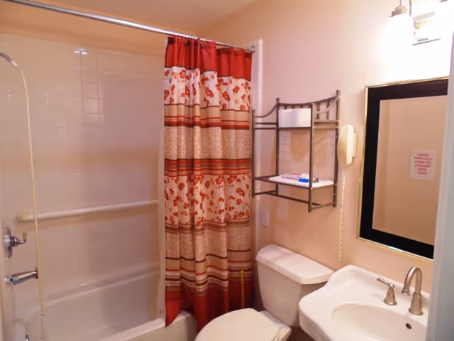 A small bathroom with a white bathtub and shower combination, a red and beige patterned shower curtain, a white toilet, a white sink with silver faucet, a wall-mounted mirror with a black frame, a wall-mounted telephone, and a metal shelf holding towels and toiletries.