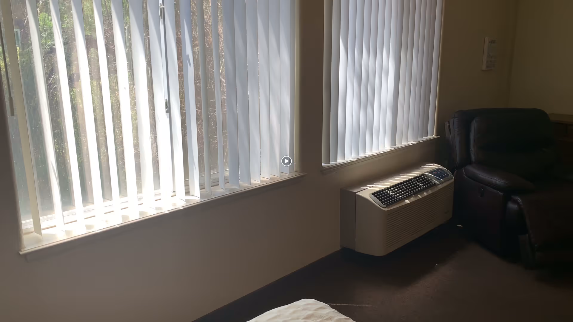 A room with two large windows covered by vertical blinds allowing sunlight to filter through. Below one window is a wall-mounted air conditioning unit. To the right, there is a dark-colored recliner chair. The floor is carpeted and the walls are light-colored.