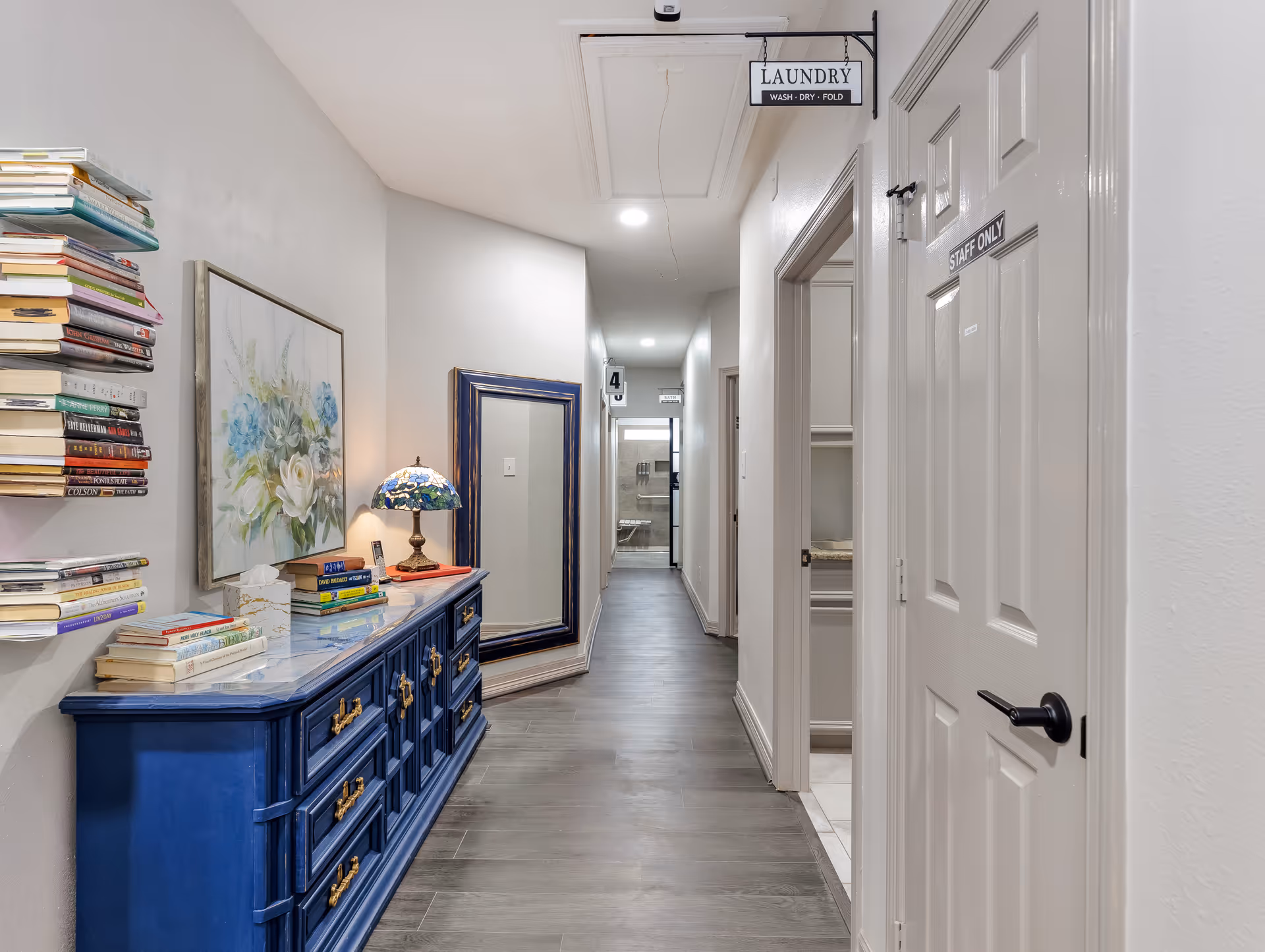 A well-lit hallway in an assisted living facility with a blue dresser topped with books and a decorative lamp on the left side. Above the dresser is a floral painting and a large mirror leaning against the wall. On the right side, there are doors including one labeled 'STAFF ONLY'. At the end of the hallway, a sign hanging from the ceiling reads 'LAUNDRY WASH DRY FOLD'. The floor is covered with gray wood-like flooring.