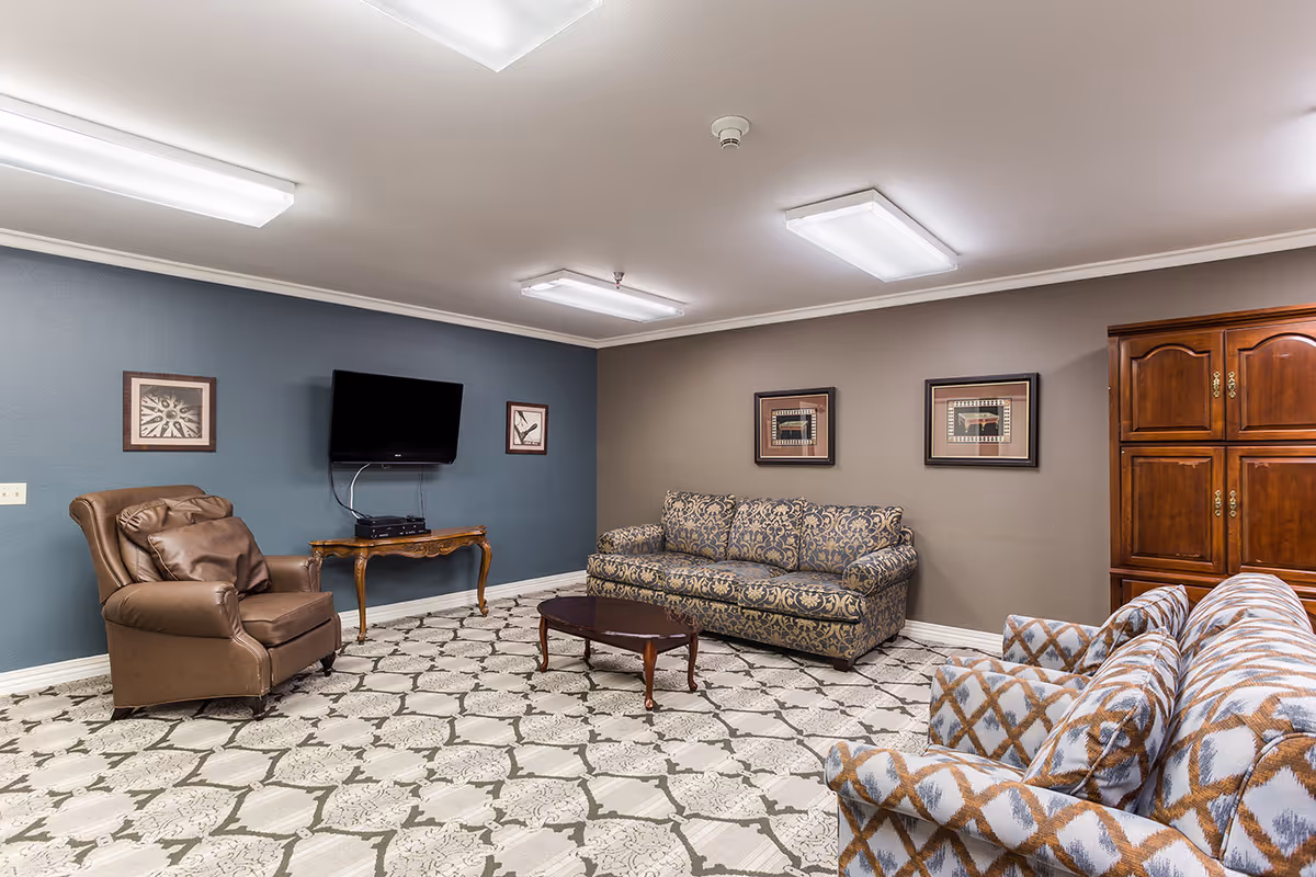 Furnished lounge with patterned sofas and armchairs, a coffee table, wall-mounted TV, framed artwork, and a wooden cabinet.