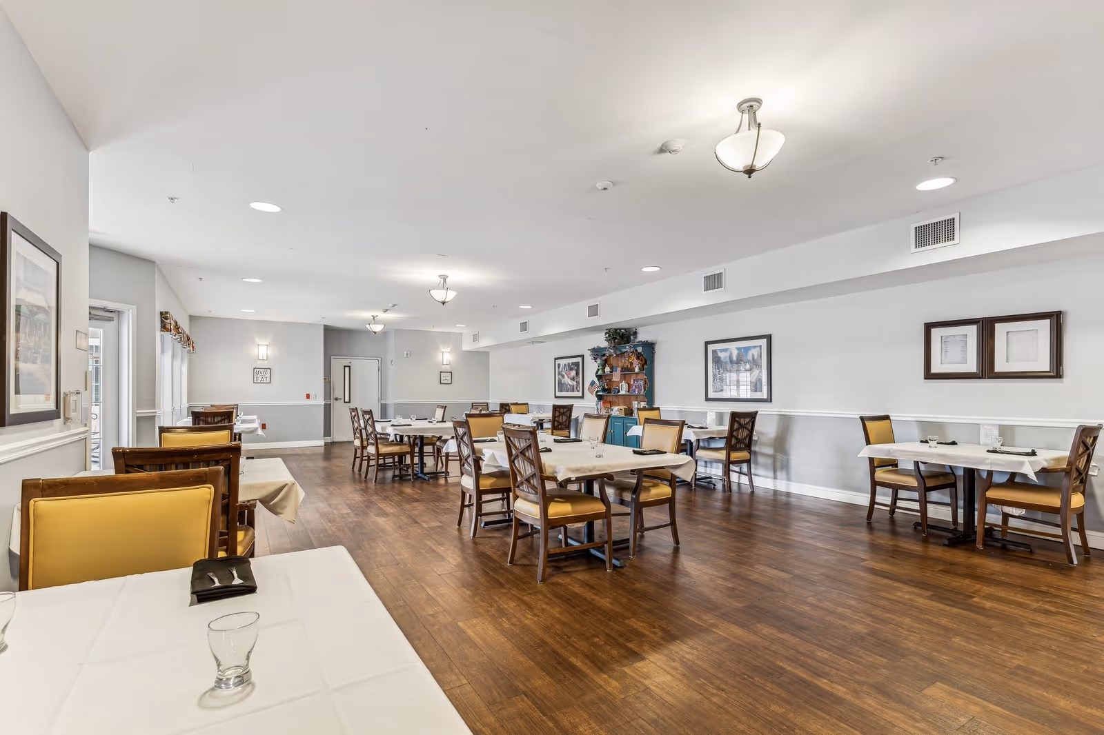 A spacious dining room with multiple tables covered with white tablecloths and surrounded by wooden chairs with yellow cushions. The room has wooden flooring, white walls with framed artwork, and ceiling lights. There is a wooden cabinet with plants and decorative items against the far wall.