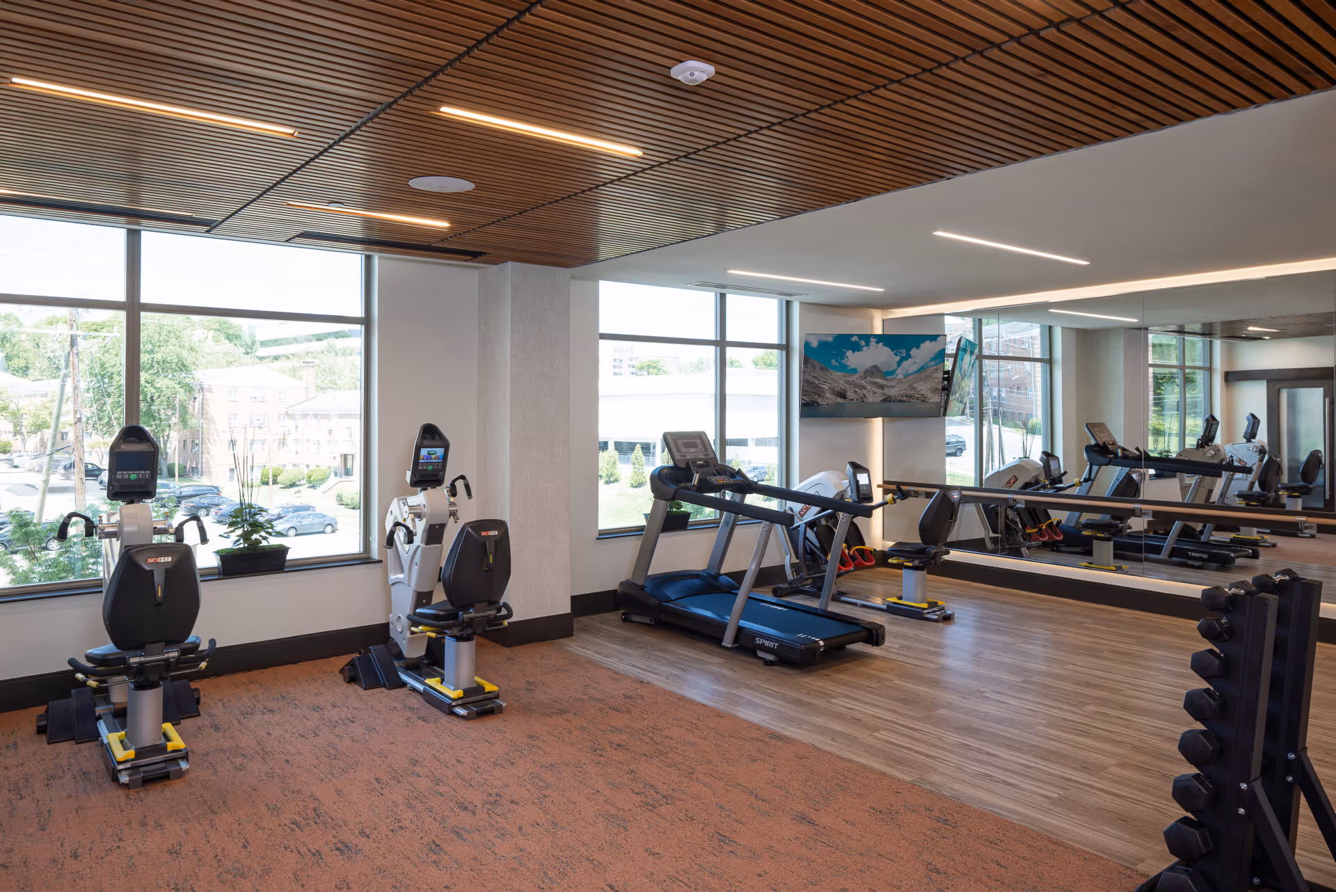 A fitness room with exercise equipment including two recumbent bikes, a treadmill, and a rack of dumbbells. The room has large windows letting in natural light, a wooden slatted ceiling with recessed lighting, and a large mirror on one wall reflecting the equipment.