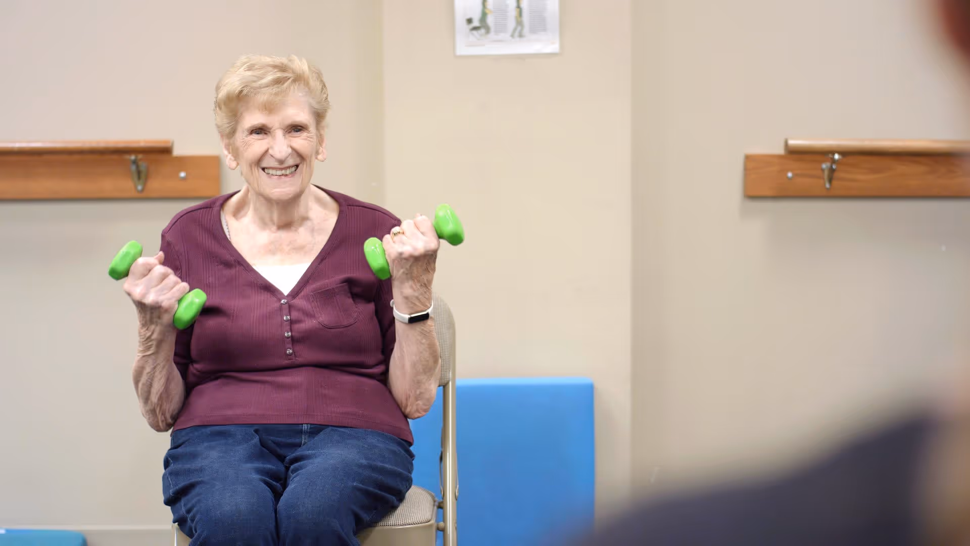 An elderly woman sitting on a chair indoors, smiling and holding a pair of small green dumbbells, engaging in light exercise.