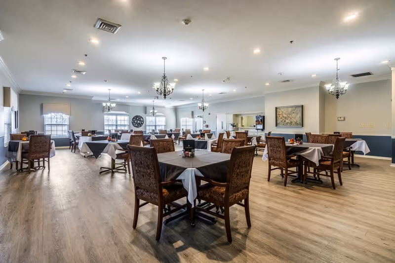 A spacious dining room with multiple tables covered with white and gray tablecloths, each surrounded by four upholstered chairs. The room has large windows allowing natural light, chandeliers hanging from the ceiling, and light wood flooring. There is a wall-mounted painting and a clock on the far wall.