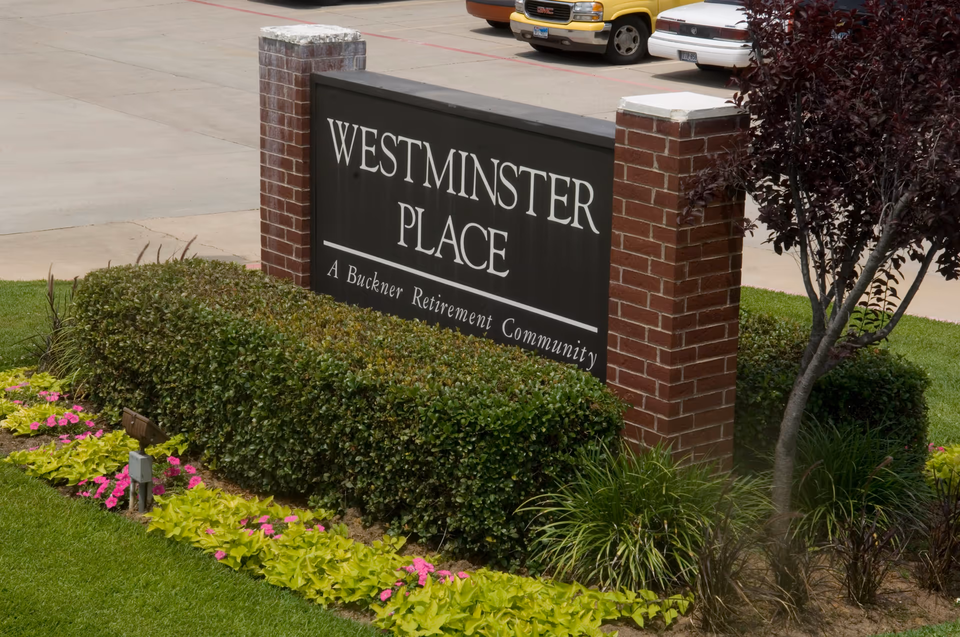 Outdoor view of a sign for Westminster Place, a Buckner Retirement Community, surrounded by neatly trimmed bushes, colorful flowers, and a small tree, with a parking lot and vehicles in the background.