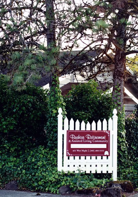 A white picket fence sign for Beehive Retirement & Assisted Living Community surrounded by green bushes and trees, with a house roof visible in the background.