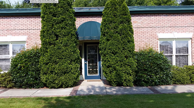 Front exterior view of a single-story brick building with a green awning over a blue door, flanked by two tall evergreen trees and surrounded by bushes and windows with white blinds.