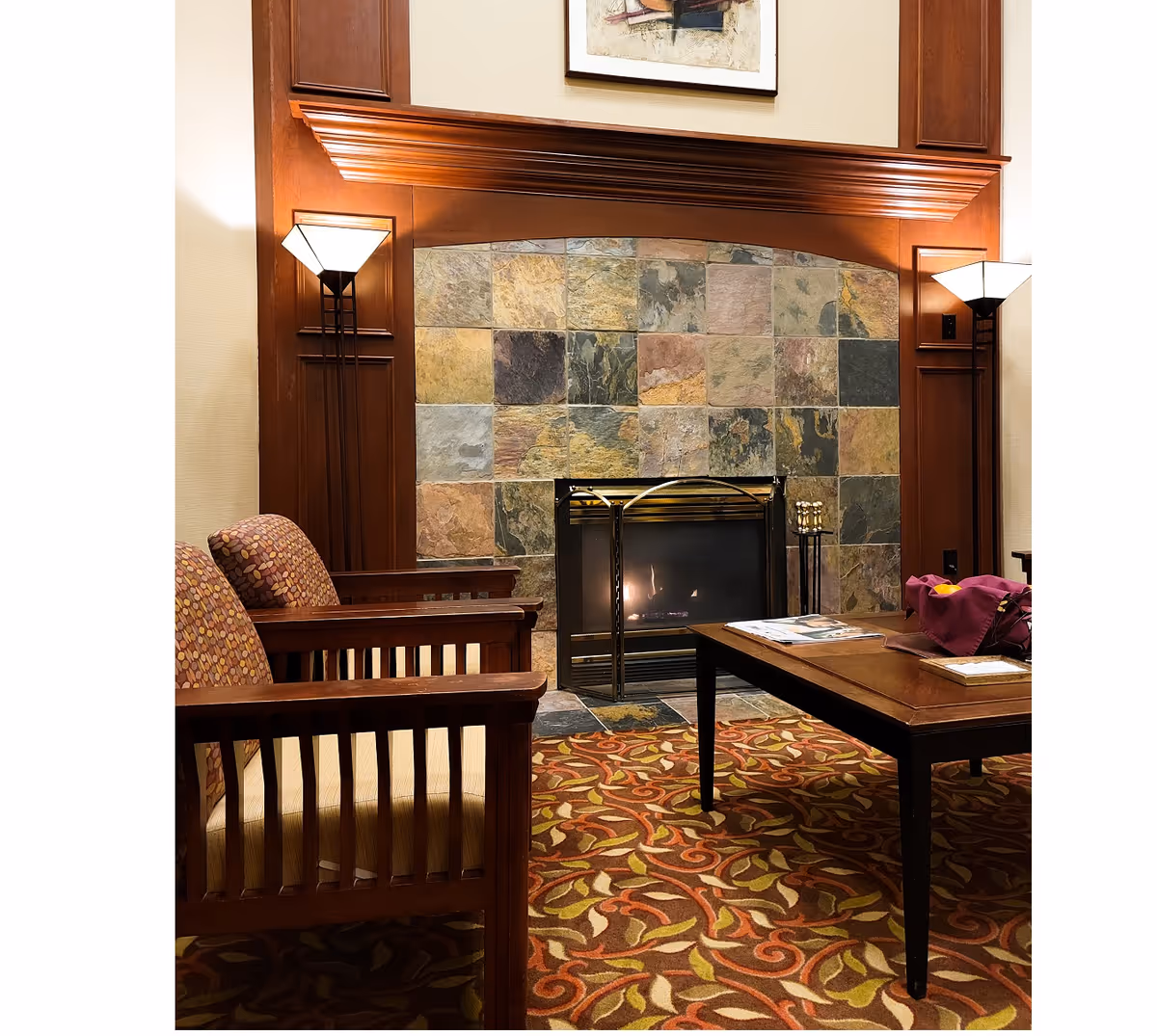 Cozy seating area with wooden chairs, patterned carpet, a stone-tiled lit fireplace, and a coffee table with magazines.