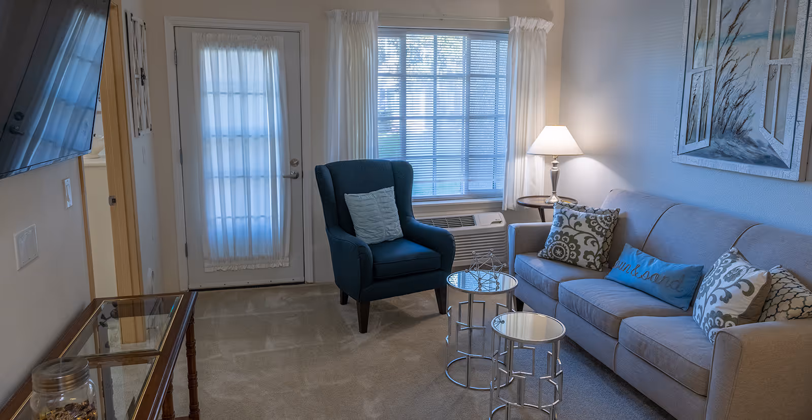 A cozy living room with a beige sofa adorned with decorative pillows, a blue armchair with a cushion, two round mirrored nesting tables, a side table with a lamp, a large window with white curtains, and a door with a sheer curtain. A flat-screen TV is mounted on the wall opposite the sofa, and a piece of artwork depicting a beach scene hangs above the sofa.