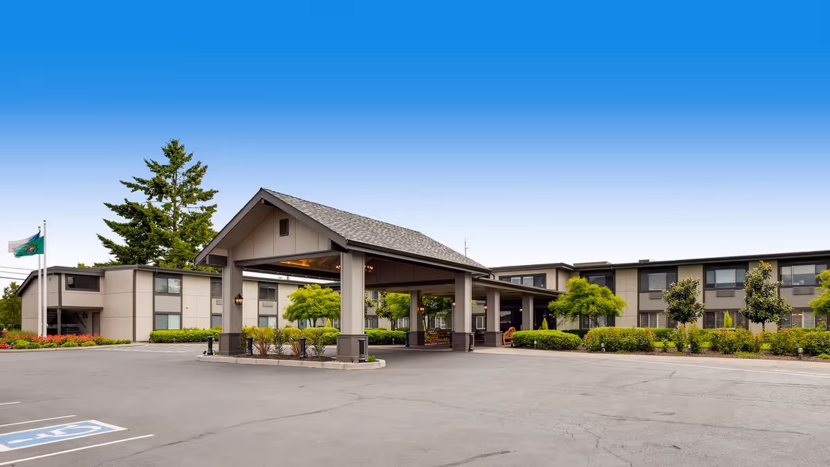 Exterior view of Cascade Inn Assisted Living Community showing a large covered entrance with a peaked roof, surrounded by a parking lot and landscaped greenery including trees and bushes under a clear blue sky.