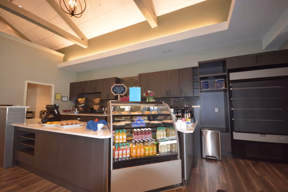 Interior view of a modern cafe or snack bar area with a display case filled with beverages and snacks, a counter with a cash register, and dark wood cabinetry in the background under a high ceiling with exposed beams and a chandelier.