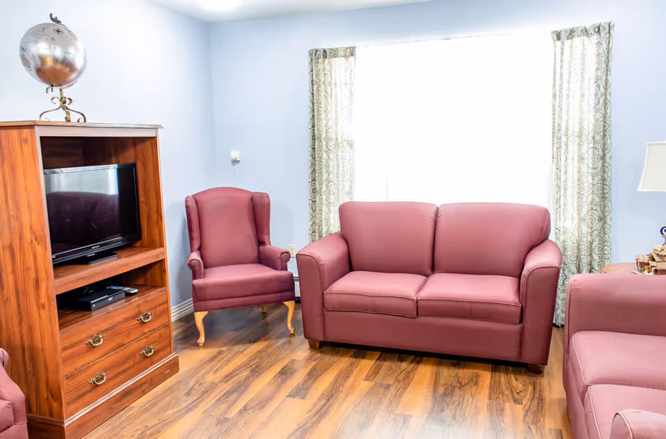 A cozy living room with wooden flooring, featuring a maroon loveseat, matching armchair, and a wooden TV stand with a flat-screen television and a decorative globe on top. Light blue walls and a large window with patterned curtains provide natural light to the room.