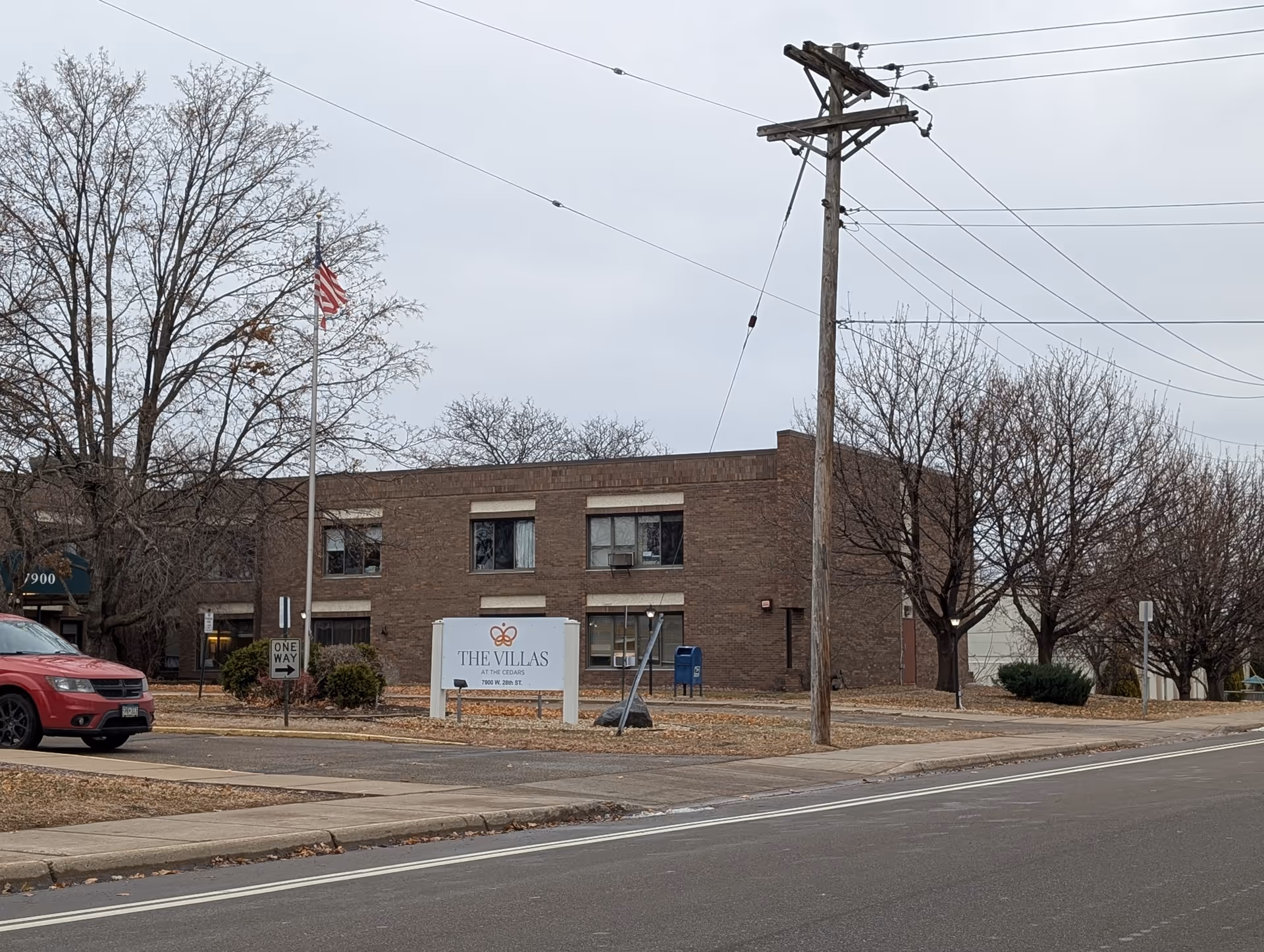 Brick two-story building with a front sign reading 'The Villas', an American flag, leafless trees, and a street in front.