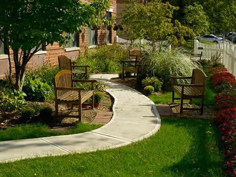A curved concrete pathway surrounded by green grass, plants, and flowers with wooden benches placed along the path next to a brick building and a white picket fence.