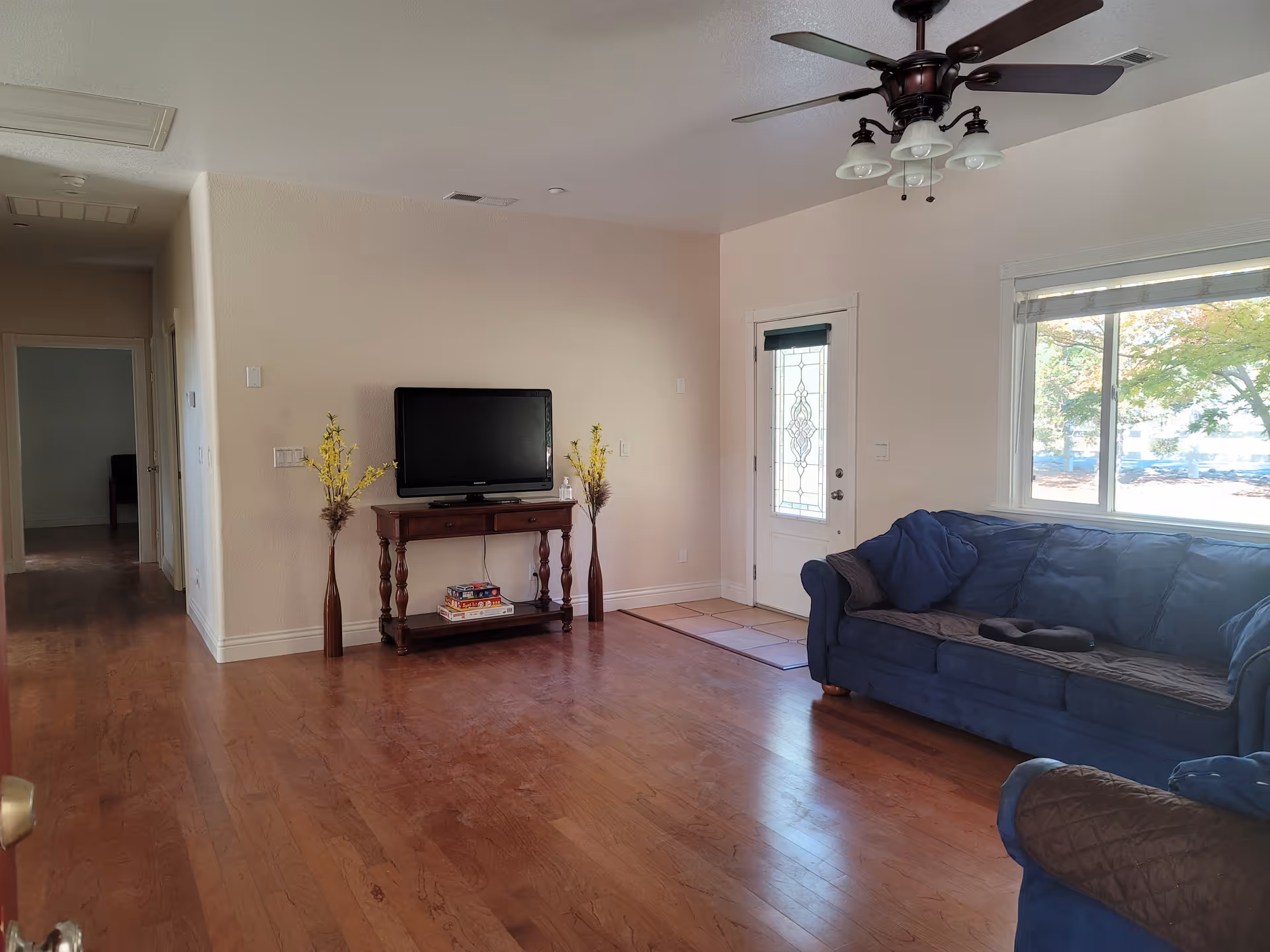 Spacious living room with hardwood floors, a blue sofa, a TV on a wooden stand, a front door and a large window.