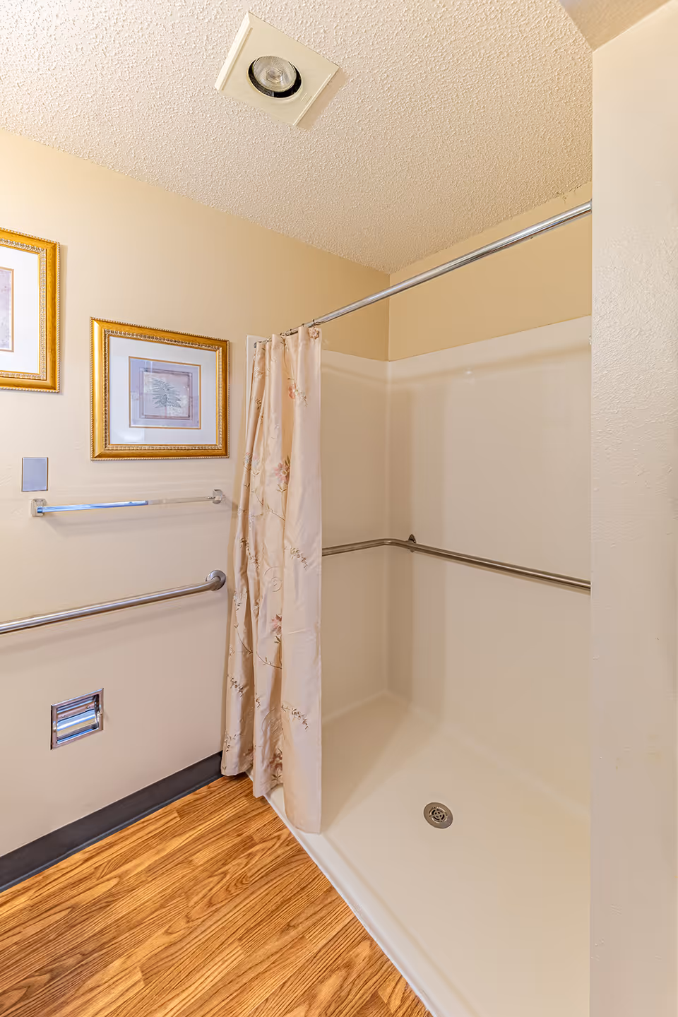 A shower area with a beige floral shower curtain partially drawn. The shower has white walls and a metal grab bar running along two sides. The floor outside the shower is wood-patterned. Two framed pictures with gold frames hang on the beige wall next to the shower, along with a towel bar and a ventilation vent near the floor.