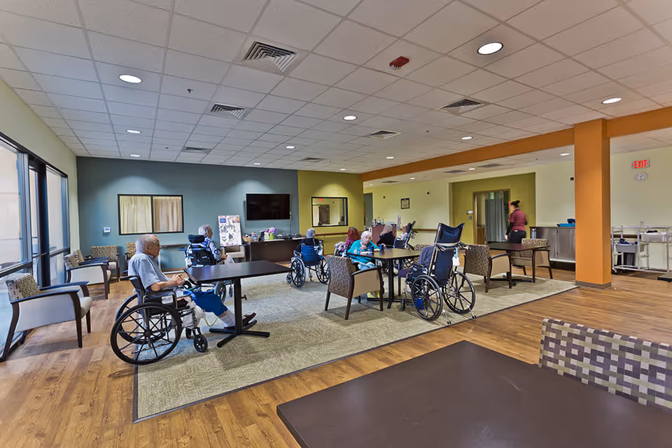 A spacious common area in a senior living facility with several elderly residents sitting in wheelchairs around tables. The room has large windows on one side, a television mounted on the wall, and a staff member walking in the background. The floor is a combination of wood and carpet, and the walls are painted in soft green and orange tones.