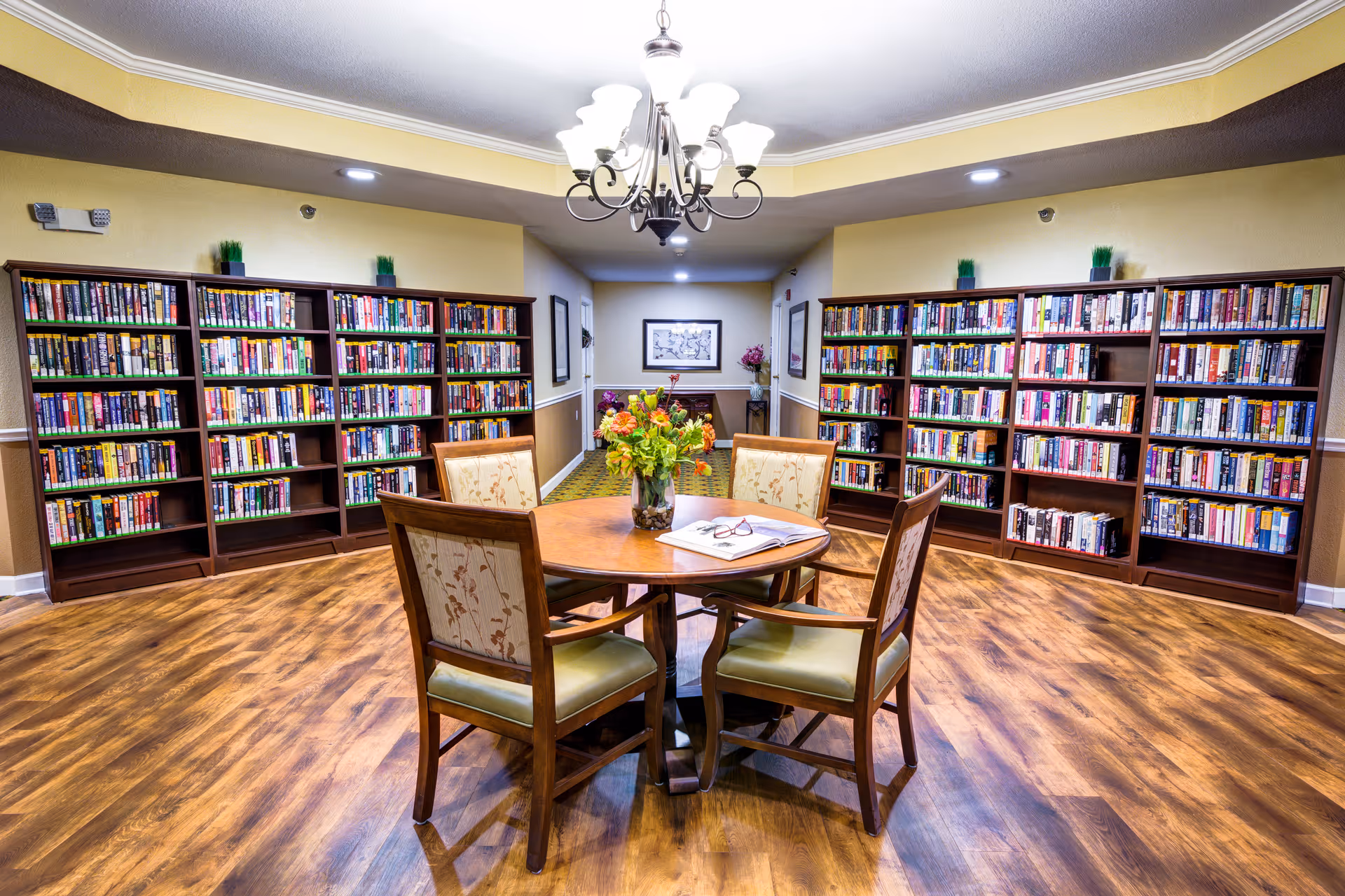 A cozy library room with wooden bookshelves filled with books lining the walls. In the center, there is a round wooden table with four cushioned chairs around it. On the table, there is a vase with flowers and an open book with reading glasses resting on it. The room has wood flooring and a chandelier hanging from the ceiling.
